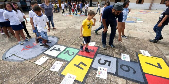 Group of children on a schoolyard playing a large colorful numbered hopscotch grid while an adult supervises nearby.
