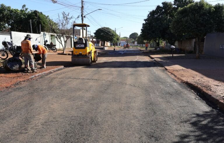 Construction workers in orange vests lay asphalt on a residential street with a yellow road roller nearby