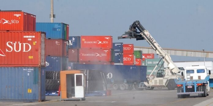 Container yard with stacked red, blue, and green shipping containers; a white reach-stacker lifts a container above the ground in the background.