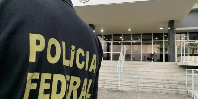 Close-up of a police officer's dark jacket with yellow 'POLICIA FEDERAL' text, in front of a public building entrance with steps and glass doors.