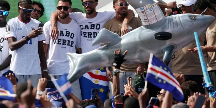 Group of men in Cabo Verde shirts pose and smile as a large shark prop is carried overhead at a beach event, with flags in the crowd foreground (Cape Verde and EU).
