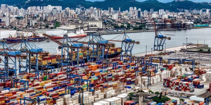 Busy container port with blue gantry cranes and colorful shipping containers by the water, city skyline and hills in the background.