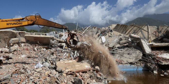 Yellow Hyundai excavator arm demolishing a concrete structure; dust plume and rubble everywhere with mountains in the distance.