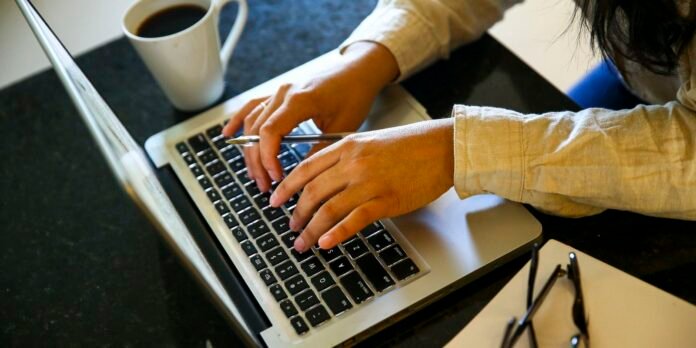 Person typing on a silver laptop at a dark desk, with a white coffee mug nearby and glasses on the side.