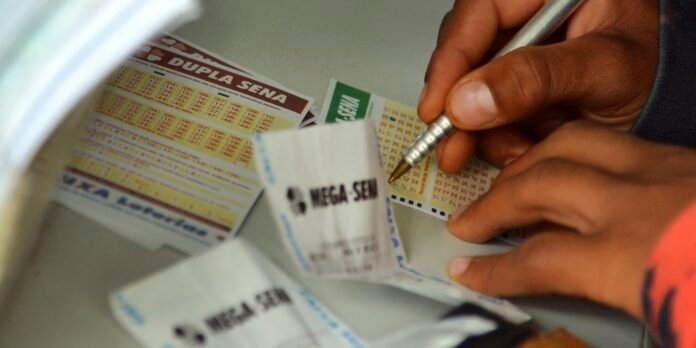 Close-up of hands writing with a pen on Mega Sena lottery tickets, with several betting slips on a table.