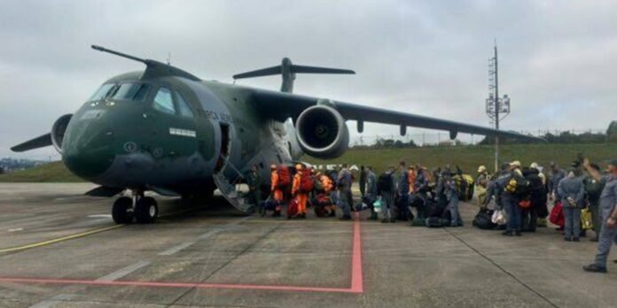 Green military transport aircraft on a tarmac with its rear ramp open and soldiers boarding or waiting to board the aircraft.
