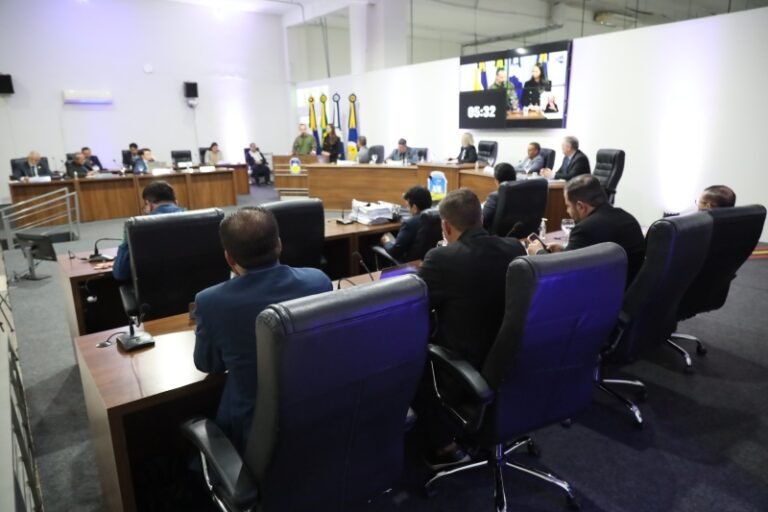 Officials seated around a large U-shaped conference table in a formal meeting room with flags and a large screen displaying a timer in the background.
