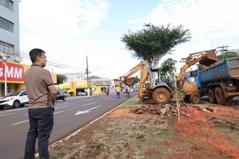 Man in a brown polo stands on a sidewalk watching construction work on a busy street lined with shops and vehicles in the background.