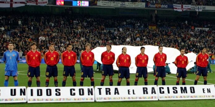 Spain soccer team lineup in red shirts on the field, goalkeeper in blue, with a banner reading 'UNIDOS CONTRA EL RACISMO EN'.