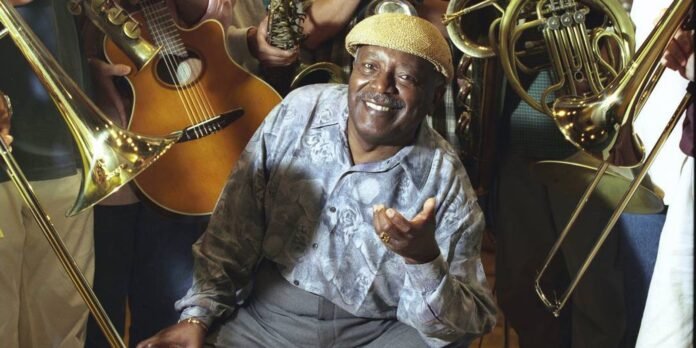 Smiling older man in a hat poses among a brass band with guitar and tubas surrounding him.
