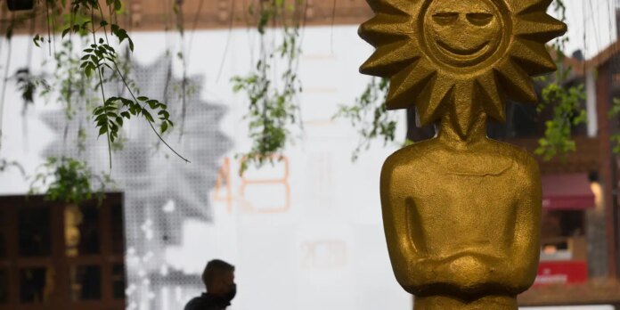 Gold sun-shaped sculpture with a smiling face atop a golden torso in a courtyard, with hanging vines overhead.