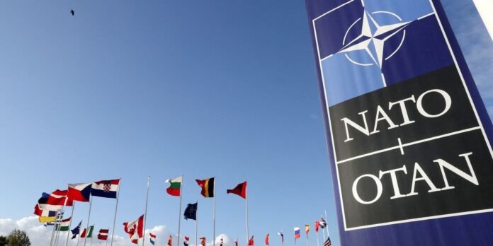 NATO flag banner on the right with a row of international flags against a clear blue sky in the background.