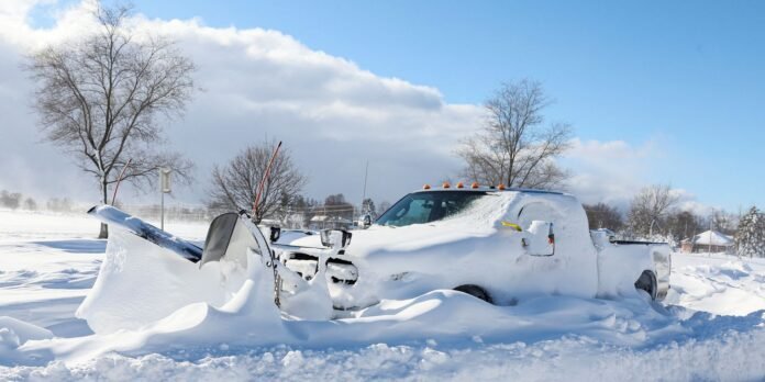 White pickup truck buried in deep snow with a snowplow blade on the left side of the image.