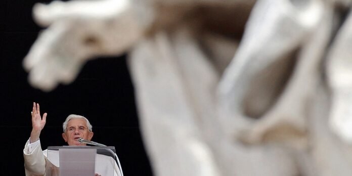 Elderly clergyman in white robes speaks at a podium with a microphone, raising his right hand, a large statue out of focus in the foreground.