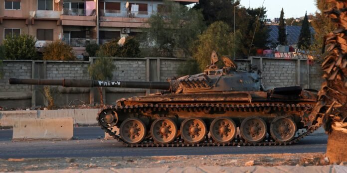 Military tank parked on an urban road beside a concrete barrier, with apartment buildings and trees in the background.