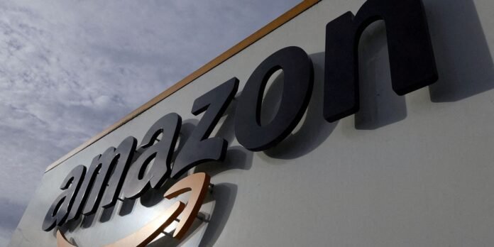 Close-up of a large outdoor sign spelling 'amazon' in bold dark letters on a white wall with an orange curved underline, angled against a cloudy sky.