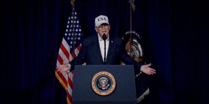 Man in a suit and a white USA cap speaks at a lectern with the presidential seal, flags in the background.