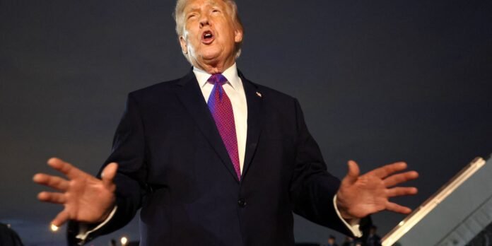 Man in a dark suit and purple tie delivers a speech with hands raised on a dim stage.
