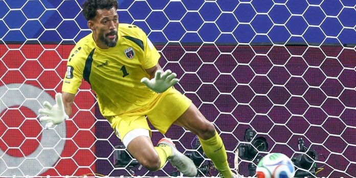 Soccer goalkeeper in yellow kit dives to his right to stop a ball near the goal, wearing white gloves and number 1 on the jersey.