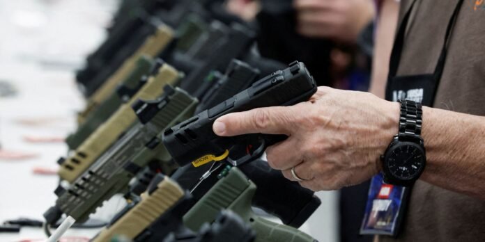 Person's hand grips a black pistol at a firearm display, with rows of other guns on a table in the background and a wristwatch visible on the handler.