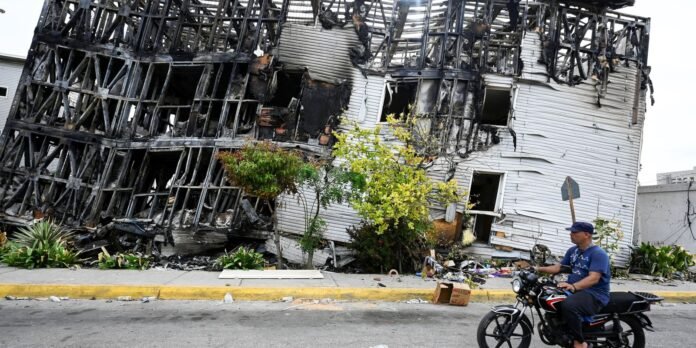 Charred, burned two-story house with exposed framing; a man on a motorcycle rides by on the street in front.