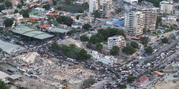 Aerial view of a city block showing collapsed buildings amid rubble, with crowds and vehicles nearby for emergency response or rescue.