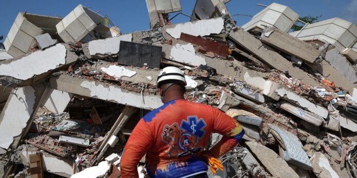 Rescue worker wearing a helmet and bright orange-blue uniform stands amid collapsed concrete and debris at a disaster site.