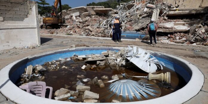 Deserted pool filled with muddy water and rubble at a collapsed building site with workers and a crane in the background.