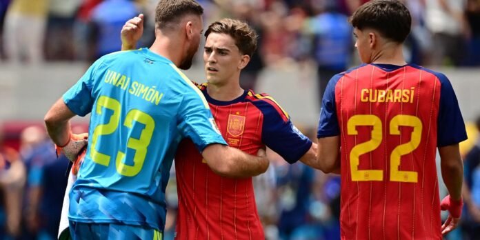 Spain national team players in red jerseys (22 Cubarši) and a blue goalkeeper (23 Unai Simón) in a crowded stadium, sharing a moment during a match.