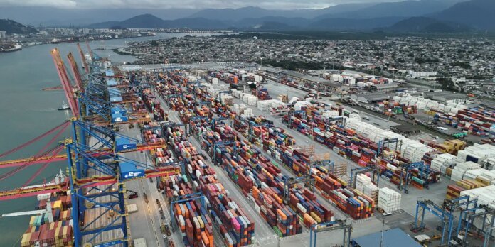 A busy container port with rows of multicolored shipping containers and towering cranes along the dock by calm water, with a city and mountains in the distance.