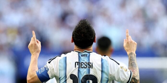 Lionel Messi seen from behind in an Argentina jersey, arms raised with middle fingers extended in a celebratory gesture.