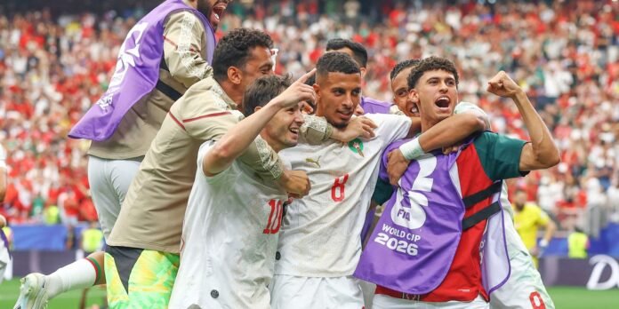 Soccer players celebrate a goal, arms around each other on the field with a cheering crowd behind them.
