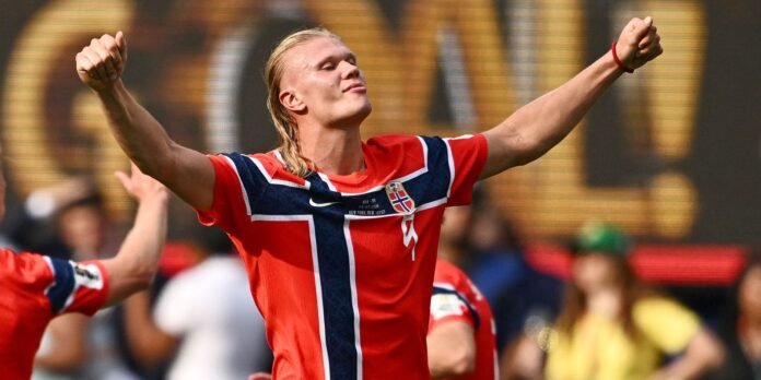 Soccer player in a red and blue Norway-inspired jersey celebrates with arms raised on the pitch.