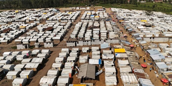 Aerial view of a large refugee camp with hundreds of white tents arranged in grids, dirt paths between them and a few colorful shelters nearby.