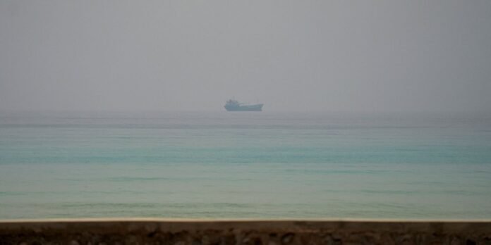Distant ship on calm turquoise sea with a hazy horizon and a railing in the foreground.