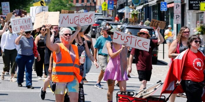 Crowd of protesters marching down a city street, holding handmade signs reading 'ICE OUT OF MAINE' and 'JUSTICE'.