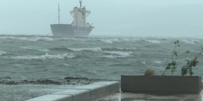 A large ship on rough seas viewed from a rocky shore with a concrete bench planter in the foreground.