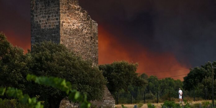 Charred stone ruin with orange flames and dark smoke in the background; a man in white observes from a fenced area.