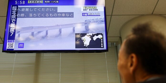Television screen in an office showing a tsunami warning news broadcast in Japanese; a person sits in the foreground watching.