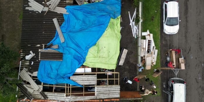Aerial view of a construction site with a partially built structure covered by blue and green tarps, corrugated metal sheets, and scattered lumber; several workers and a white van parked nearby along a street.