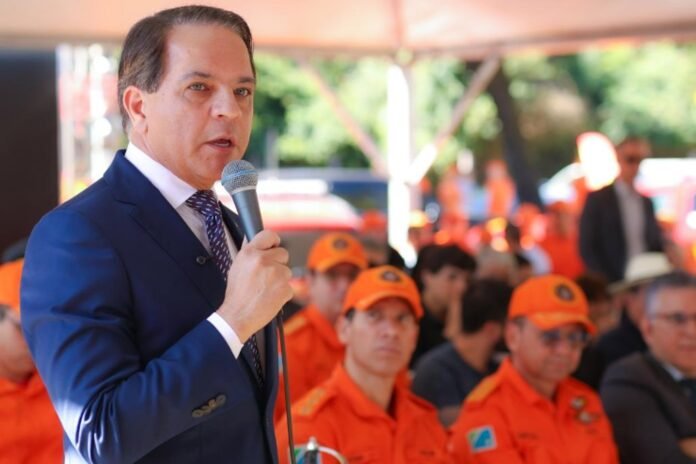 Man in a navy suit speaks into a microphone at an outdoor event, with orange-uniformed attendees behind him.