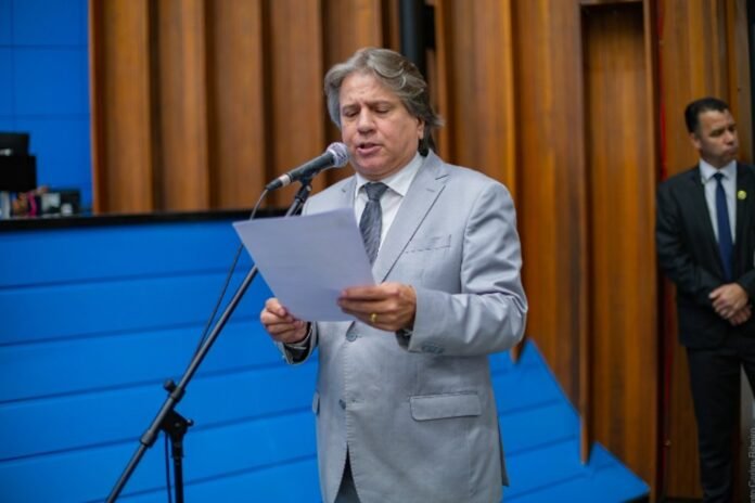 Man in a light gray suit reads from a sheet while speaking into a microphone at a podium, with a blue wall and wooden panels behind him and a security guard in the background.
