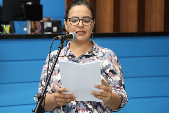 Woman with glasses reads from a sheet at a microphone on a blue stage backdrop in a studio setting.