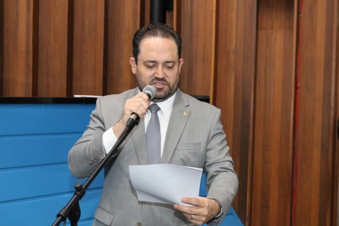 Man in a gray suit speaks into a microphone while reading from papers, in a wood-paneled room.