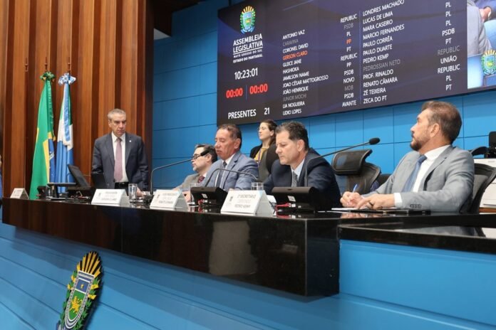 Panel of lawmakers seated at a curved desk with microphones, nameplates, and flags in a government chamber.