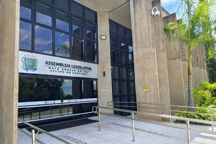 Front entrance of a government building with a sign reading 'Assembleia Legislativa' and glass doors, a metal ramp, and palm trees nearby.
