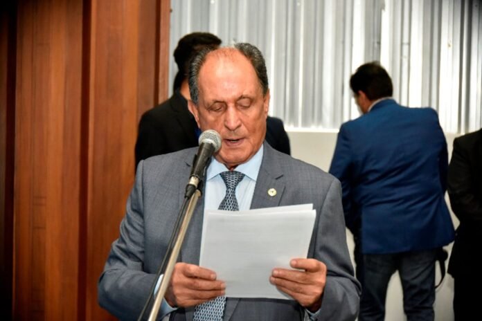 Older man in a gray suit reads from papers at a microphone during a formal event.