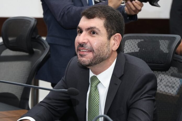 Man in a dark suit and green tie sits at a conference table with a microphone in front, speaking or preparing to speak in a meeting room.