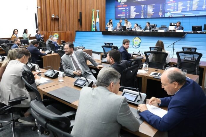 Parliamentary chamber with officials seated at desks, a large blue wall and flags in the background, and a digital board displaying numbers above.
