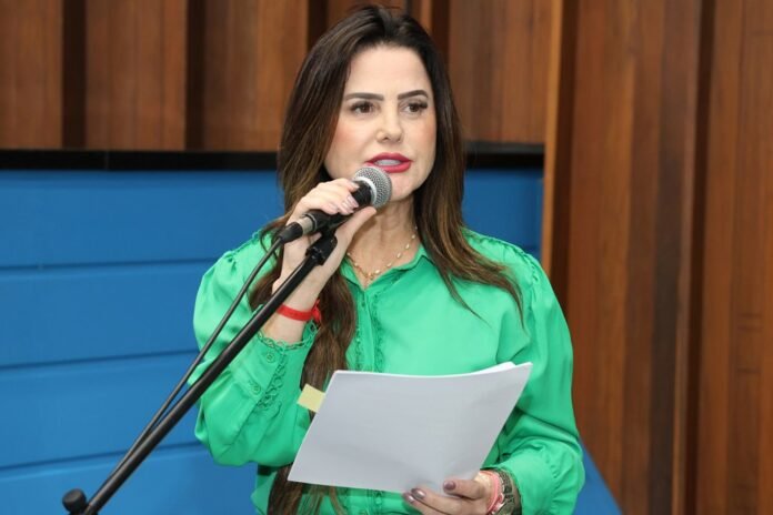 Woman in a bright green blouse speaks into a handheld microphone while holding papers at a podium or microphone stand.